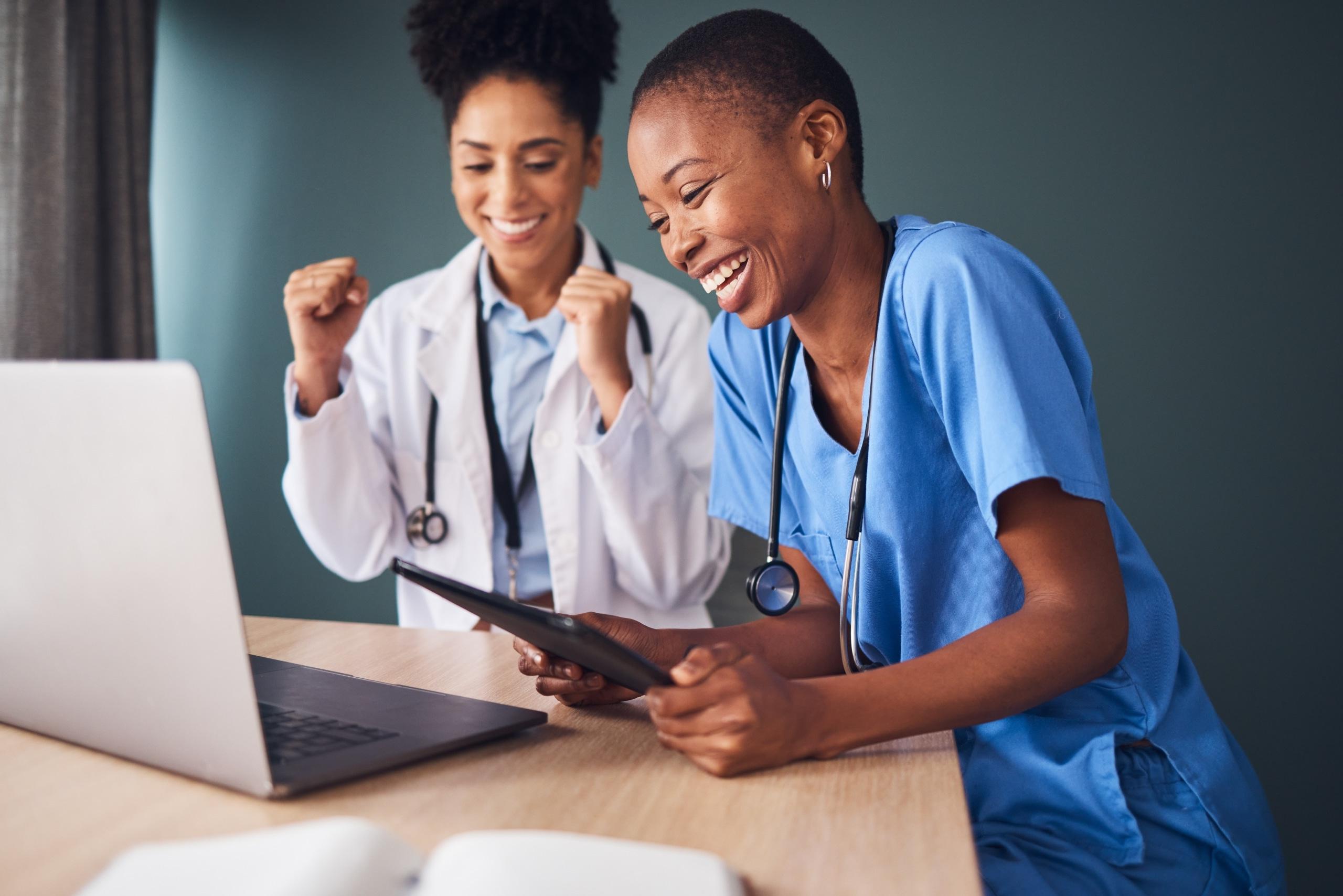 Two nurses laughing together as they use a tablet and laptop